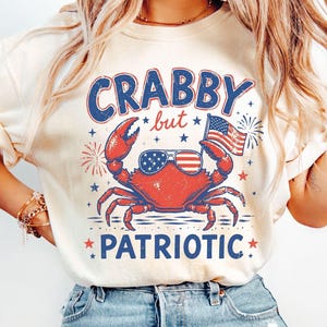 Op de afbeelding: Ivoor kleurig T-shirt met een afbeelding van een rode krab met een zonnebril en een Amerikaanse vlag. De tekst "Crabby but Patriotic" is in blauw en rood. Het ontwerp bevat vuurwerk en sterren.