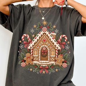 Op de afbeelding: Een antracietgrijs t-shirt met korte mouwen en een centrale afbeelding van een peperkoekhuisje, omringd door twee peperkoekmannetjes, zuurstokken, hulstbladeren en sneeuwvlokken. Het ontwerp bevat rode en witte accenten en sterren.