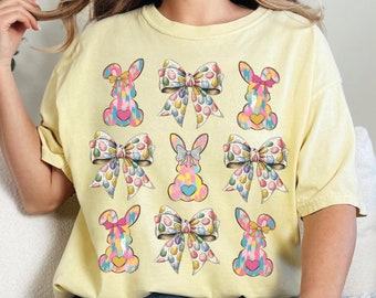 May include: A yellow t-shirt featuring a design of colourful Easter bunnies and bows. The bunnies are wearing bows with pastel colours and candy-like designs.