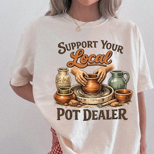 May include: Ivory-colored t-shirt with the text "Support Your Local Pot Dealer" in brown and orange. The graphic features hands shaping clay on a pottery wheel, surrounded by pottery. The shirt is a relaxed fit.