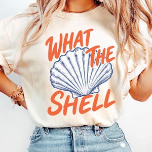 May include: Ivory-colored t-shirt with the phrase "WHAT THE SHELL" in orange and a blue and white seashell graphic. The shirt is paired with light blue denim jeans.