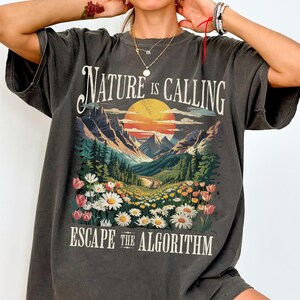 May include: Dark gray t-shirt with a scenic graphic. The design features a sunset over mountains, a road, and flowers, with the text "NATURE IS CALLING" and "ESCAPE THE ALGORITHM" in white.