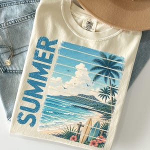 May include: Ivory-colored t-shirt with a beach scene graphic. The design features a blue ocean, palm trees, and surfboards, with the word "SUMMER" in blue. A tan hat and blue jeans are also in the image.