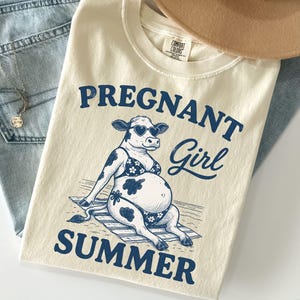 Könnte beinhalten: Elfenbeinfarbenes T-Shirt mit dem navyblauen Schriftzug "PREGNANT Girl SUMMER". Die Grafik zeigt eine Kuh mit herzförmiger Sonnenbrille und Bikini. Ein beiger Hut und blaue Jeans sind ebenfalls im Bild zu sehen.