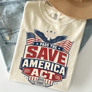Peut inclure: Un t-shirt ivoire avec une impression graphique représentant un pygargue à tête blanche, deux drapeaux américains et le Capitole des États-Unis. Le texte indique : PASS THE SAVE AMERICA ACT. Le motif utilise une palette de rouge, blanc et bleu.