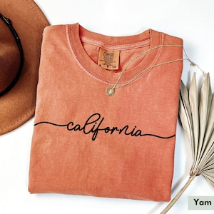 May include: A peach-colored t-shirt with the word "california" written in a cursive font across the front. The shirt is folded and laid flat on a white surface.
