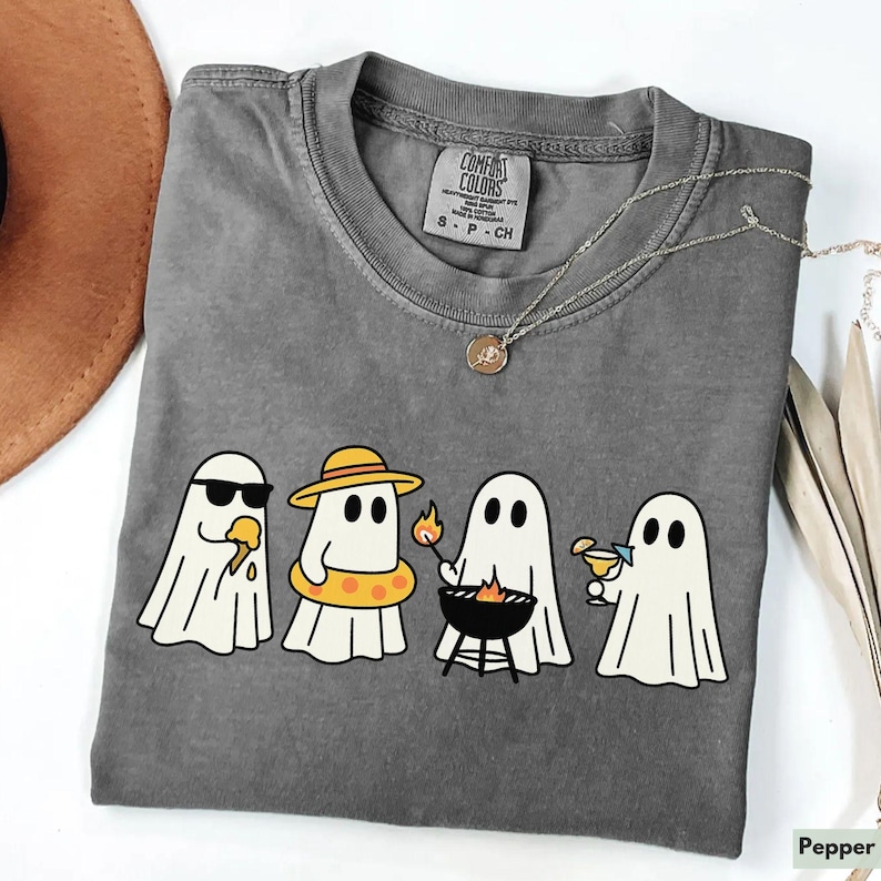 May include: A gray Comfort Colors t-shirt featuring a graphic of four cartoon ghosts enjoying summer activities. One ghost wears sunglasses and eats ice cream, another wears a hat and floatie, one grills, and the last sips a cocktail.
