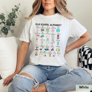 May include: White t-shirt with a graphic design of an alphabet with retro icons. Each letter of the alphabet is paired with a word and a retro icon, such as 'Aa Arcade', 'Bb Boom Box', 'Cc Cassette', 'Dd Disco Ball', 'Ee Etch A Sketch', 'Ff Floppy Disc', 'Gg Groovy', 'Hh Hula Hoop', 'Ii Ice Cream Parlor', 'Jj Jukebox', 'Kk Kite', 'Ll Lava Lamp', 'Mm Music', 'Nn Neon', 'Oo Overalls', 'Pp Polaroid', 'Qq Queen', 'Rr Record Player', 'Ss Skates', 'Tt Television', 'Uu Unique', 'Vv Vhs', 'Ww Walkman', 'Xx Xylophone', 'Yy Yo-Yo', 'Zz Zig Zag'.