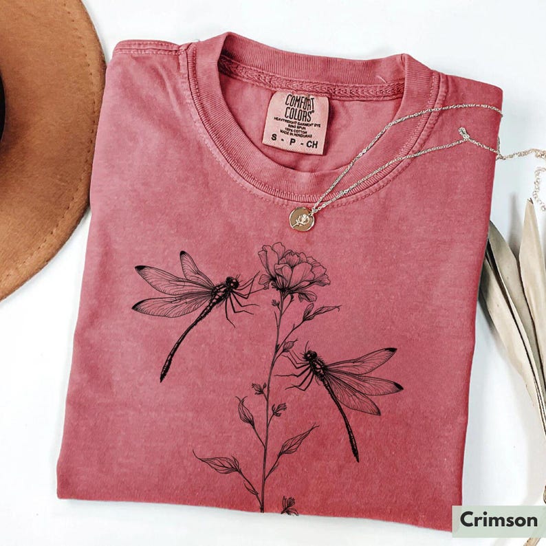 May include: A crimson red t-shirt with a floral design featuring two dragonflies and the text "COMFORT COLORS" on a white tag. The tag also includes the text "S - P - CH".