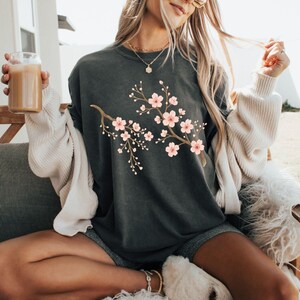 May include: A dark gray oversized t-shirt with a pink and brown floral design. The shirt is worn by a person with long blonde hair and sunglasses. The person is sitting on a white furry chair.