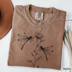 May include: A brown t-shirt with a floral design and two dragonflies. The shirt has the text "COMFORT COLORS" and "S - P - CH" printed on the tag.