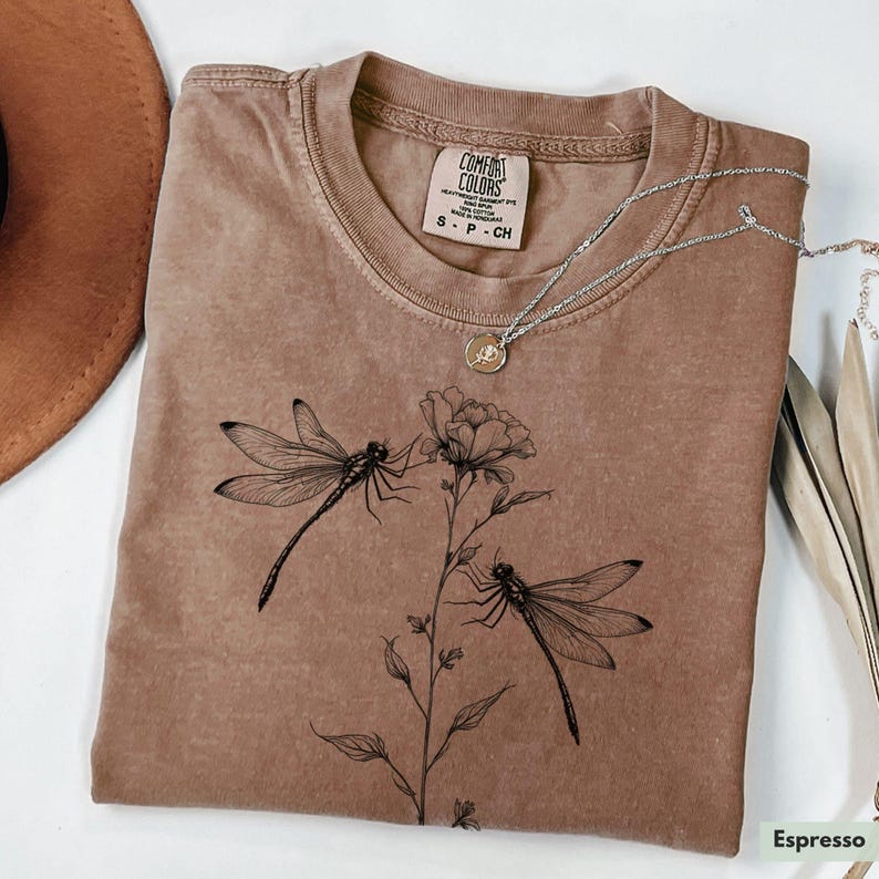 May include: A brown t-shirt with a floral design and two dragonflies. The shirt has the text "COMFORT COLORS" and "S - P - CH" printed on the tag.