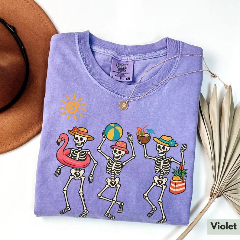 May include: A purple t-shirt with a graphic of three skeletons wearing summer hats and holding beach items. The skeletons are wearing sunglasses and are smiling. The text "Comfort Colors" is on a tag attached to the shirt.