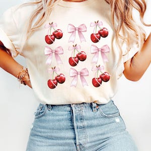 May include: A white t-shirt with a pattern of red cherries and pink bows. The shirt is made of a soft, comfortable material and has a relaxed fit.