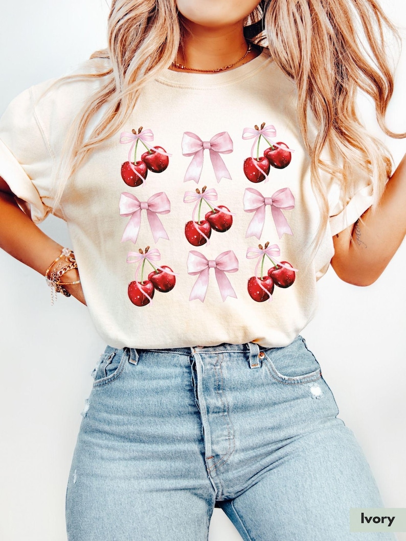 May include: A white t-shirt with a pattern of red cherries and pink bows. The shirt is made of a soft, comfortable material and has a relaxed fit.