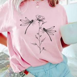 May include: A pink t-shirt with a black and white graphic of two dragonflies and a flower.
