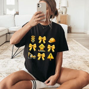 May include: Black t-shirt with a graphic print of yellow construction signs, a traffic light, and bows. The signs include "Under Construction" and an exclamation point.