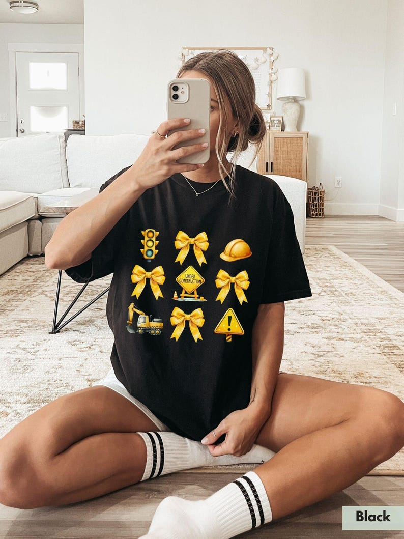 May include: Black t-shirt with a graphic print of yellow construction signs, a traffic light, and bows. The signs include "Under Construction" and an exclamation point.