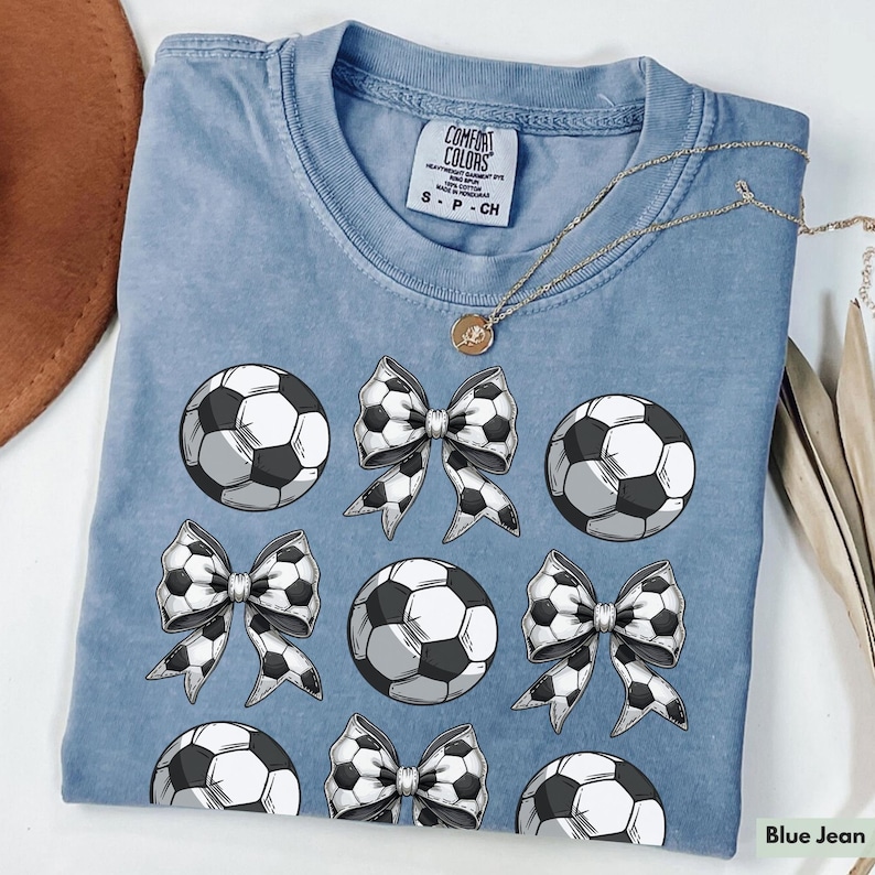 Puede incluir: Una camiseta azul vaquero con un patr&oacute;n de bal&oacute;n de f&uacute;tbol blanco y lazo. La camiseta tiene el texto "Comfort Colors" en la etiqueta.