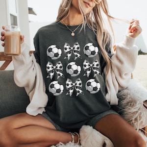 Puede incluir: Una sudadera gris oscuro con un dise&ntilde;o gr&aacute;fico de balones de f&uacute;tbol blanco y negro y lazos. La sudadera est&aacute; siendo usada por una persona.