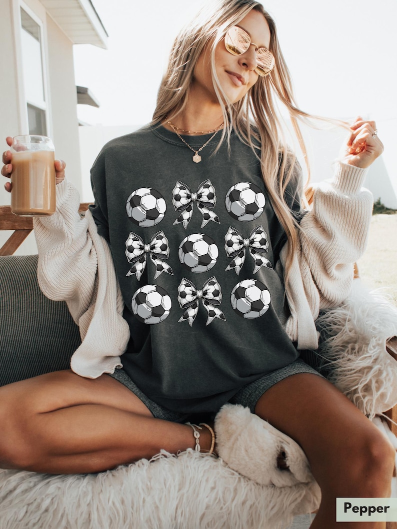 Puede incluir: Una sudadera gris oscuro con un dise&ntilde;o gr&aacute;fico de balones de f&uacute;tbol blanco y negro y lazos. La sudadera est&aacute; siendo usada por una persona.