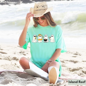 May include: A light turquoise t-shirt featuring a graphic of four cartoon ghosts enjoying summer activities. One wears sunglasses, another a sun hat and inflatable ring, one grills, and the last holds a cocktail. The shirt is worn on a beach.