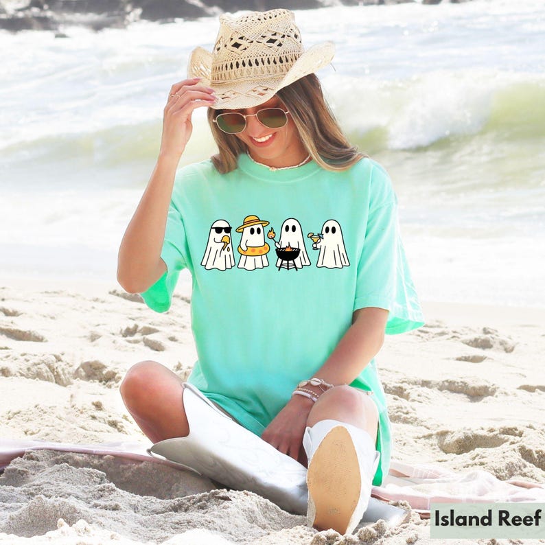 May include: A light turquoise t-shirt featuring a graphic of four cartoon ghosts enjoying summer activities. One wears sunglasses, another a sun hat and inflatable ring, one grills, and the last holds a cocktail. The shirt is worn on a beach.