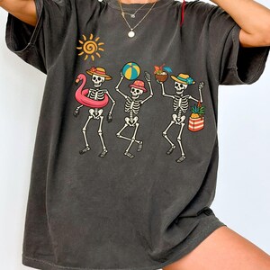 May include: A dark gray oversized t-shirt with a graphic print of three skeletons enjoying summer activities. The skeletons are wearing sunglasses and holding a beach ball, a flamingo float, and a cocktail.