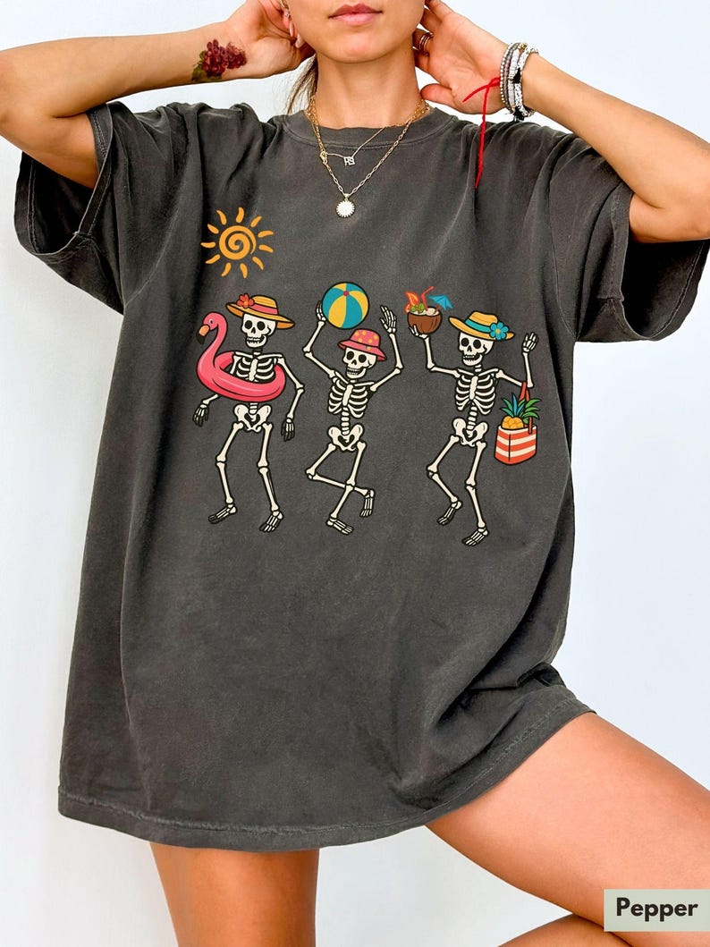 May include: A dark gray oversized t-shirt with a graphic print of three skeletons enjoying summer activities. The skeletons are wearing sunglasses and holding a beach ball, a flamingo float, and a cocktail.