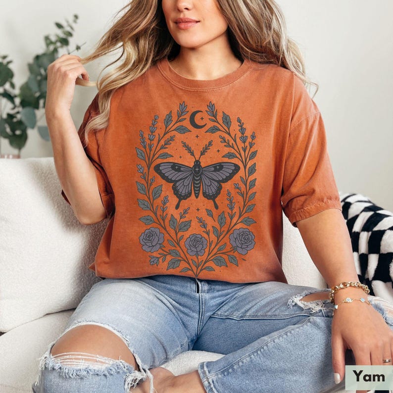 May include: A rust-colored t-shirt with a graphic of a butterfly in a floral wreath. The butterfly is dark gray and the flowers are white and gray.