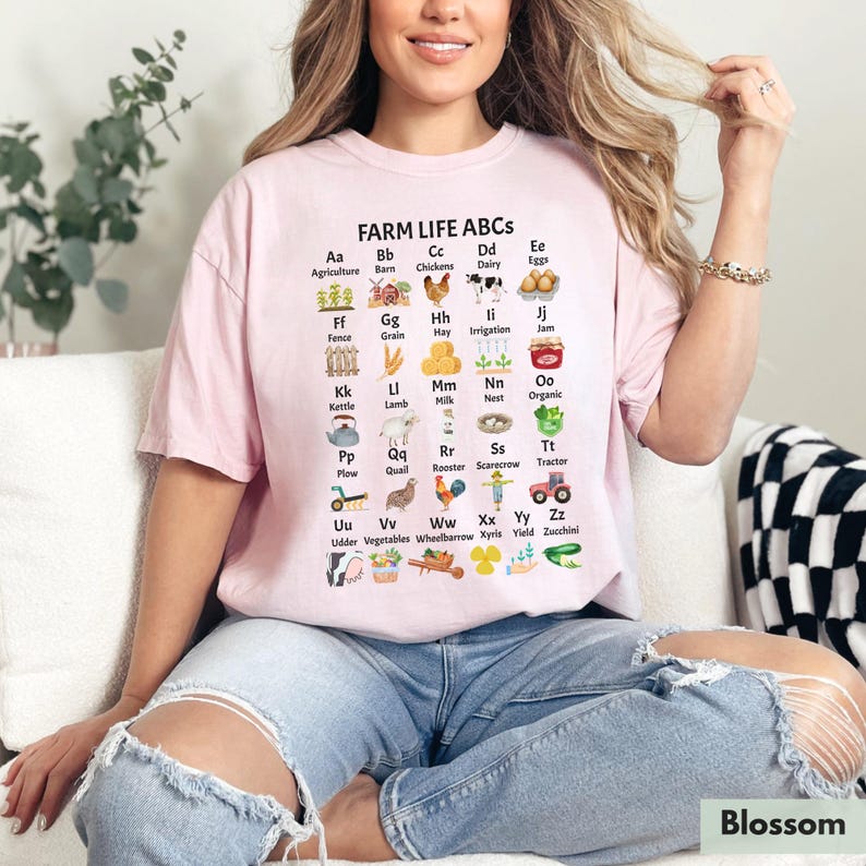 May include: A pink t-shirt featuring a white graphic of farm-related items and the alphabet. The graphic says "Farm Life ABCs" and includes images of a barn, chickens, dairy, eggs, fence, grain, hay, irrigation, jam, kettle, lamb, milk, nest, organic, plough, quail, rooster, scarecrow, tractor, udder, vegetables, wheelbarrow, xyris, yield, and zucchini.