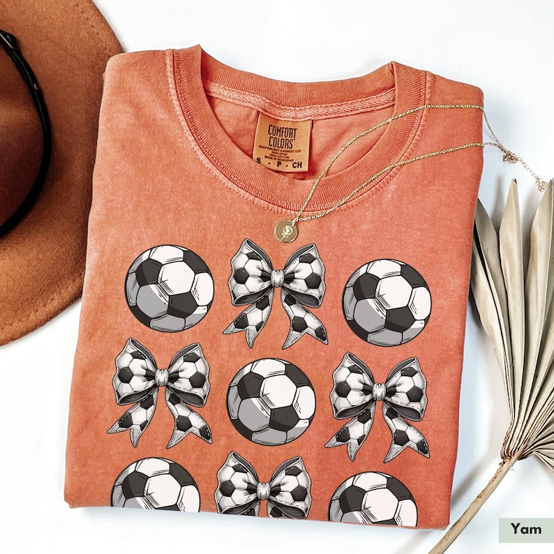 Puede incluir: Una camiseta de color coral con un dise&ntilde;o gr&aacute;fico de balones de f&uacute;tbol blanco y negro y lazos. La camiseta tiene una etiqueta que dice "Comfort Colors".