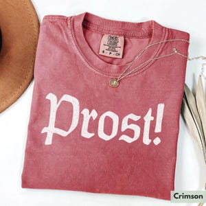 Puede incluir: Una camiseta de color rojo carmes&iacute; con la palabra "Prost!" impresa en blanco en la parte delantera. La camiseta est&aacute; hecha de una tela suave y c&oacute;moda y tiene un aspecto vintage y desgastado.