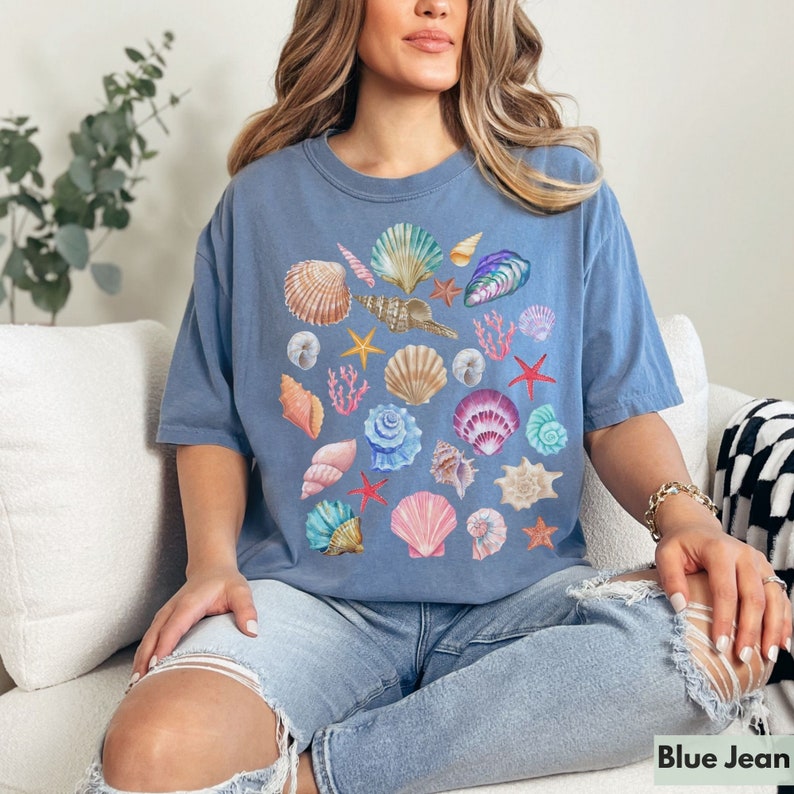Op de afbeelding: Een blauw T-shirt met een kleurrijke schelpenprint. Het shirt toont verschillende schelpen, zeesterren en koralen in tinten roze, paars, oranje en geel.