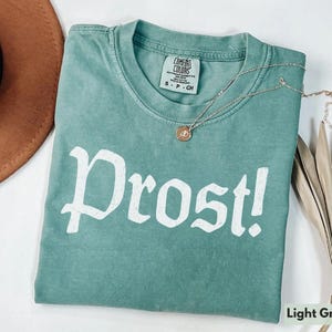 Puede incluir: Camiseta verde claro con la palabra "Prost!" impresa en blanco en la parte delantera. La camiseta est&aacute; hecha de un material suave y c&oacute;modo y tiene un corte holgado.