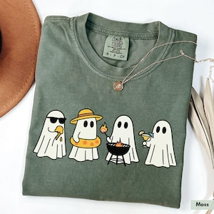 May include: A moss green Comfort Colors t-shirt featuring a graphic of four cartoon ghosts enjoying summer activities. One wears sunglasses and eats ice cream, another wears a hat and floatie, one grills, and the last one sips a cocktail.
