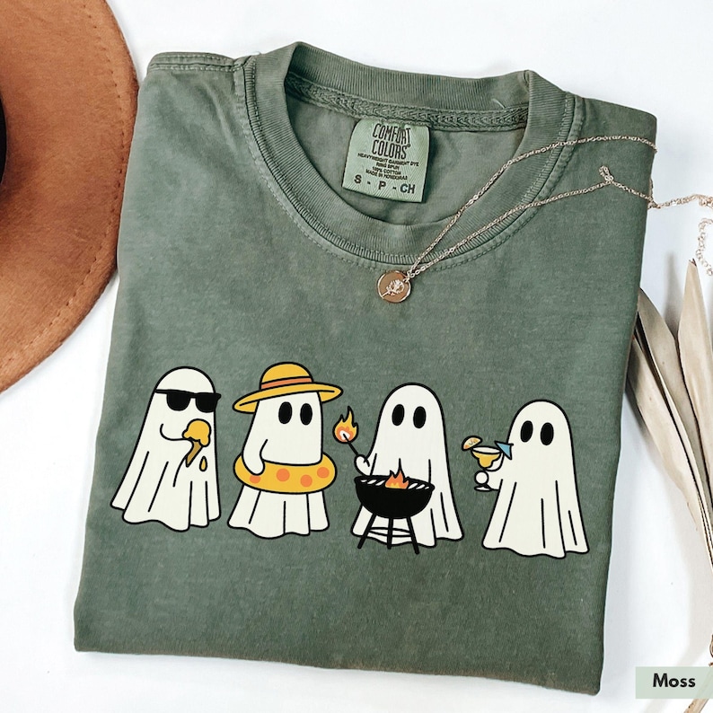 May include: A moss green Comfort Colors t-shirt featuring a graphic of four cartoon ghosts enjoying summer activities. One wears sunglasses and eats ice cream, another wears a hat and floatie, one grills, and the last one sips a cocktail.