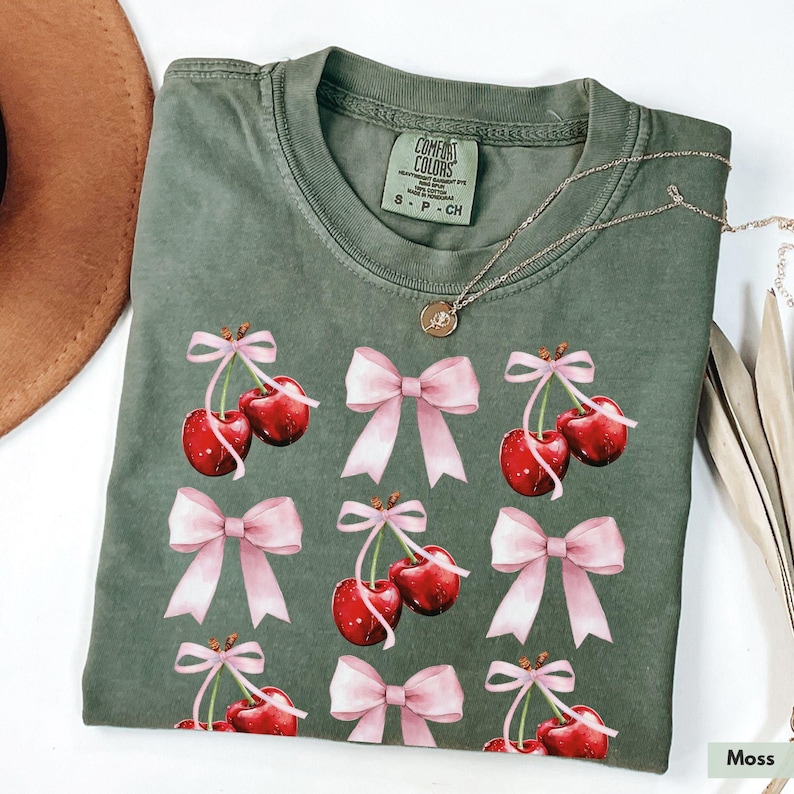 May include: A green t-shirt with a pink bow and cherry print. The shirt has the text "Comfort Colors" and "S - P - CH" on the tag.