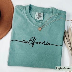 May include: Light green t-shirt with the word "california" written in black cursive script. The shirt has a distressed look and is made of a soft, comfortable fabric.