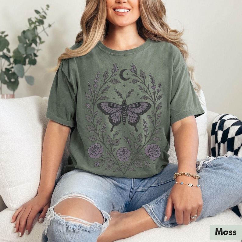 May include: A green t-shirt with a floral design featuring a black butterfly and a crescent moon. The design is in shades of purple and black.