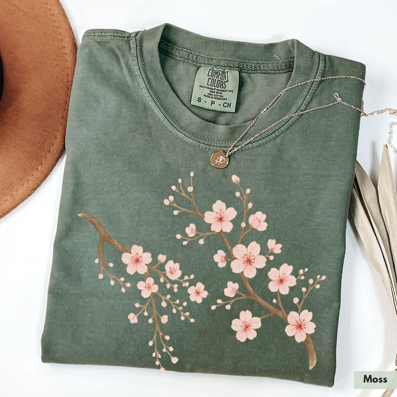 May include: A moss green t-shirt with a pink cherry blossom design. The shirt has a crew neck and short sleeves. The text "COMFORT COLORS" is printed on a tag attached to the shirt.