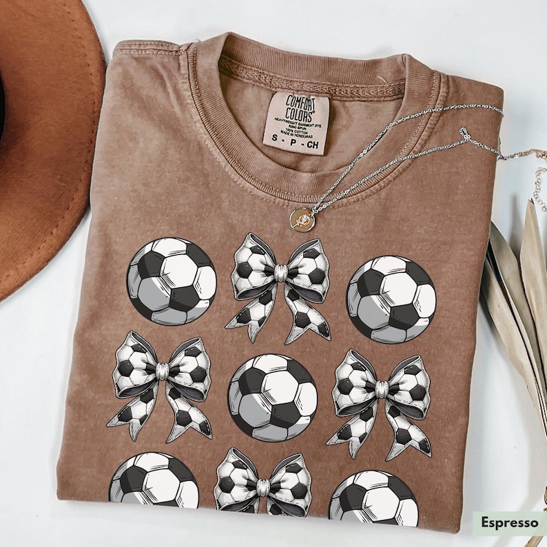 Puede incluir: Una camiseta marr&oacute;n con un patr&oacute;n de bal&oacute;n de f&uacute;tbol y lazos. La camiseta tiene el texto "Comfort Colors" y "S - P - CH" en la etiqueta. El color de la camiseta es "Espresso".