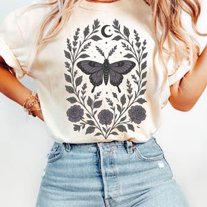 May include: A cream colored t-shirt with a graphic of a butterfly surrounded by floral vines and a crescent moon. The butterfly is black and purple with a white outline. The floral vines are black and gray with purple flowers.