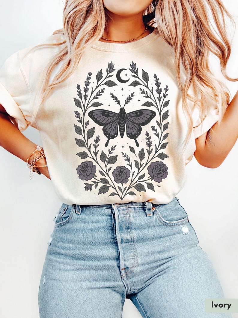May include: A cream colored t-shirt with a graphic of a butterfly surrounded by floral vines and a crescent moon. The butterfly is black and purple with a white outline. The floral vines are black and gray with purple flowers.