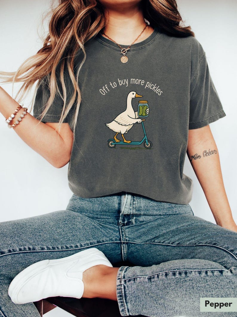 May include: Dark grey t-shirt featuring a white goose on a blue scooter, carrying a pickle jar. The text reads "off to buy more pickles". The shirt is made of a soft material. A fun, casual design.