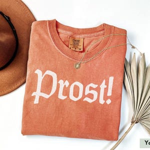 May include: A coral colored t-shirt with the word "Prost!" printed in white on the front. The shirt is made of a soft, comfortable fabric and has a relaxed fit.