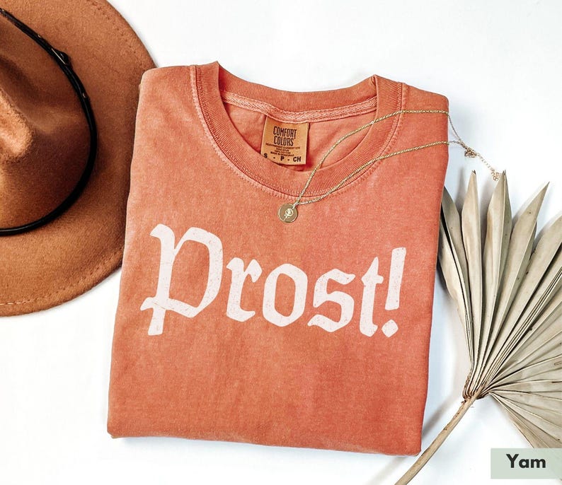 Puede incluir: Una camiseta de color coral con la palabra "Prost!" impresa en blanco en la parte delantera. La camiseta est&aacute; hecha de una tela suave y c&oacute;moda y tiene un corte holgado.
