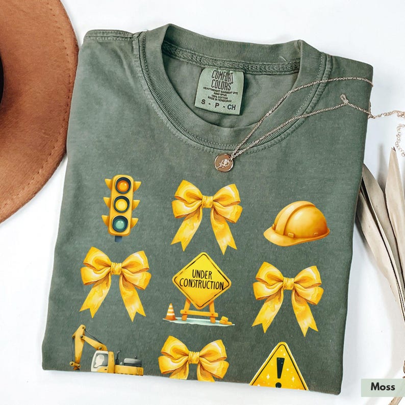 May include: A green t-shirt with yellow construction-themed graphics. The graphics include a traffic light, a construction sign that says "Under Construction", a hard hat, and bows. The shirt is made by Comfort Colors.