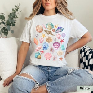 Op de afbeelding: Wit T-shirt met een kleurrijk schelpdesign. Het shirt heeft een verscheidenheid aan schelpen in verschillende vormen, maten en kleuren, waaronder roze, blauw, paars en wit.