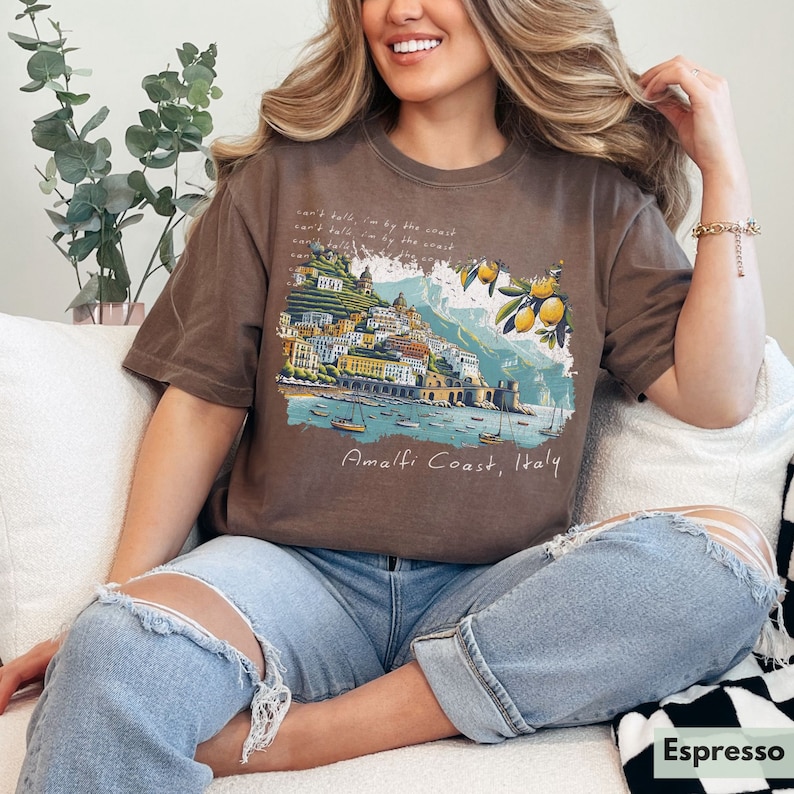 以下が含まれることがあります： イタリアのアマルフィ海岸のグラフィックプリントが施されたブラウンのTシャツ。グラフィックには、カラフルな街並みと水に浮かぶボート、レモンの木が描かれています。グラフィックの下には「Amalfi Coast, Italy」の文字がプリントされています。